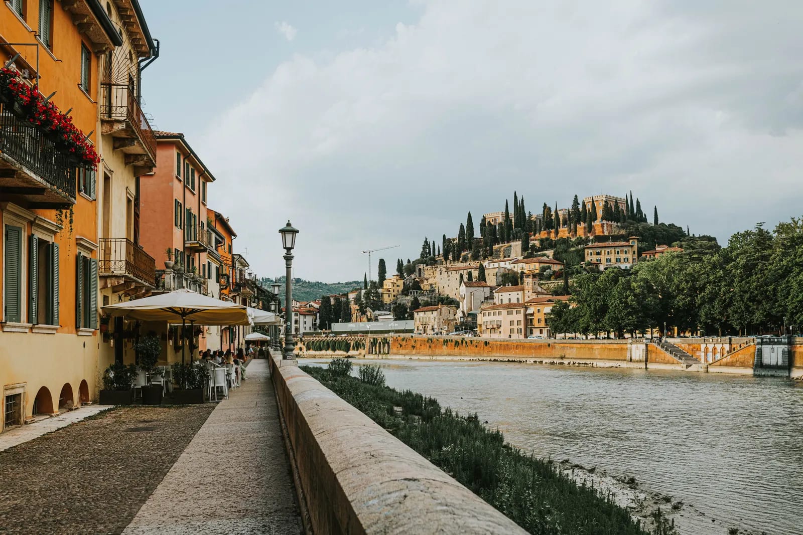 Paseo junto al río Adigio en Verona, con edificios de fachadas ocres, una terraza con sombrillas y la colina del Castel San Pietro al fondo
