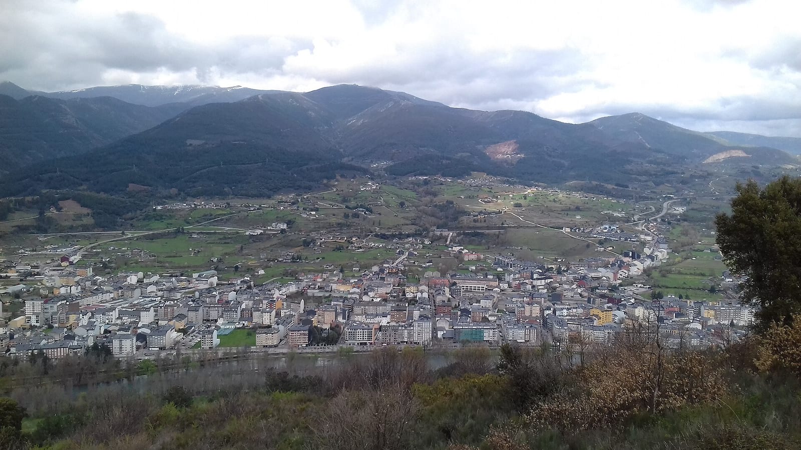 Vista desde un monte de un pueblo extendido en un valle rodeado de montañas boscosas, bajo un cielo cubierto de nubes