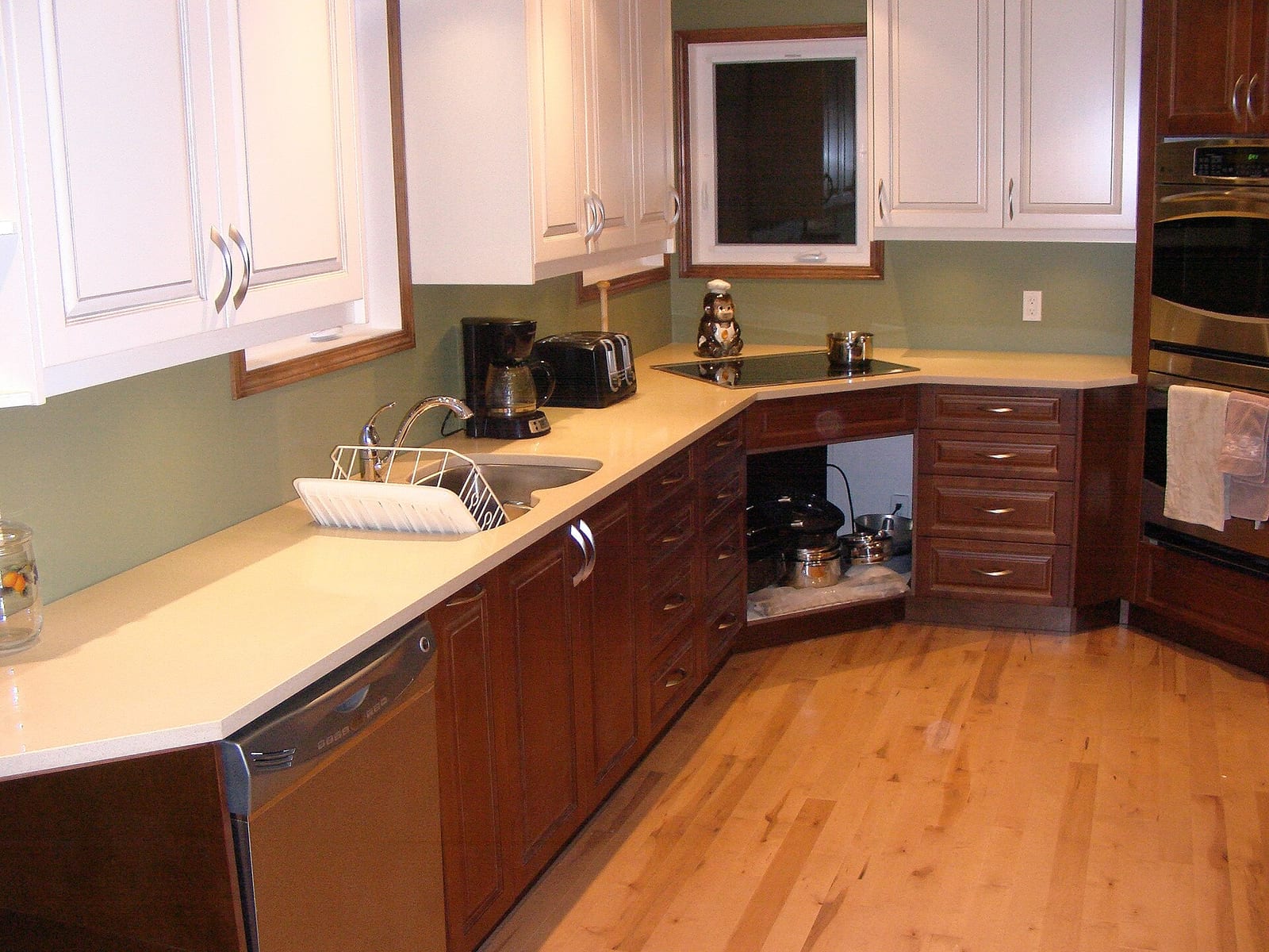 Cocina en L con encimera clara de piedra artificial, fregadero de acero con escurridor, armarios de madera y placa de cocción