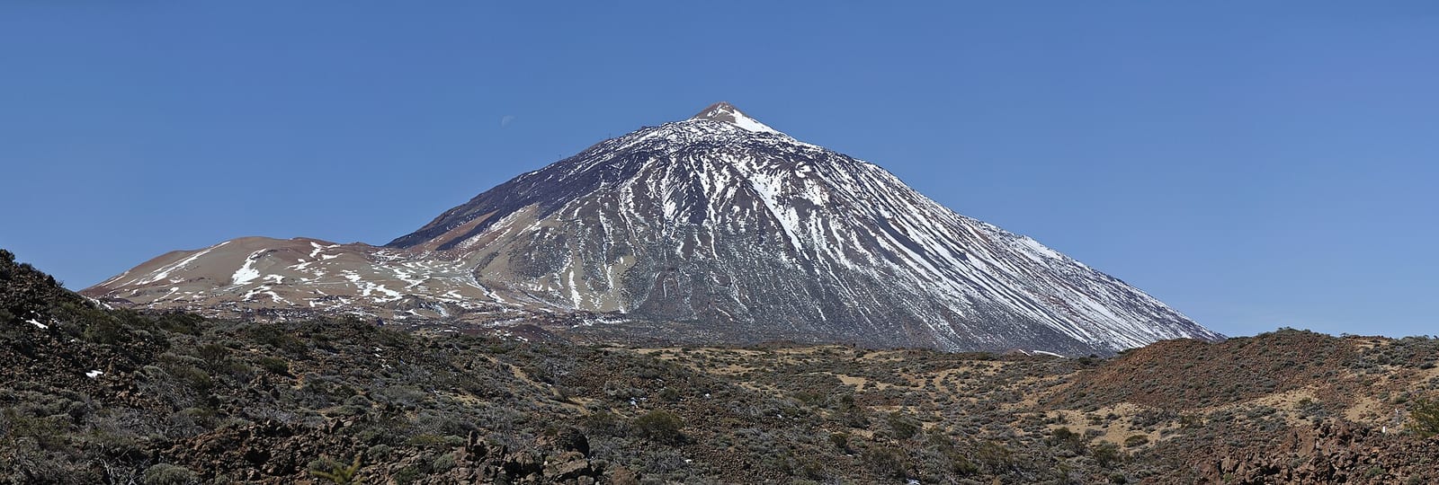 Cono volcánico del Teide con la cima nevada bajo un cielo despejado, con vegetación baja y terreno volcánico en primer plano