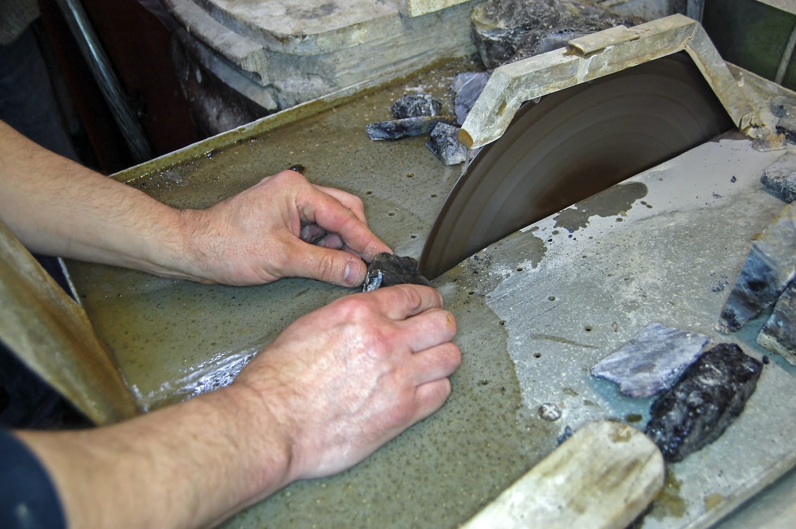 Dos manos sujetan un fragmento de mineral junto al disco de una sierra circular refrigerada por agua, con virutas de roca alrededor