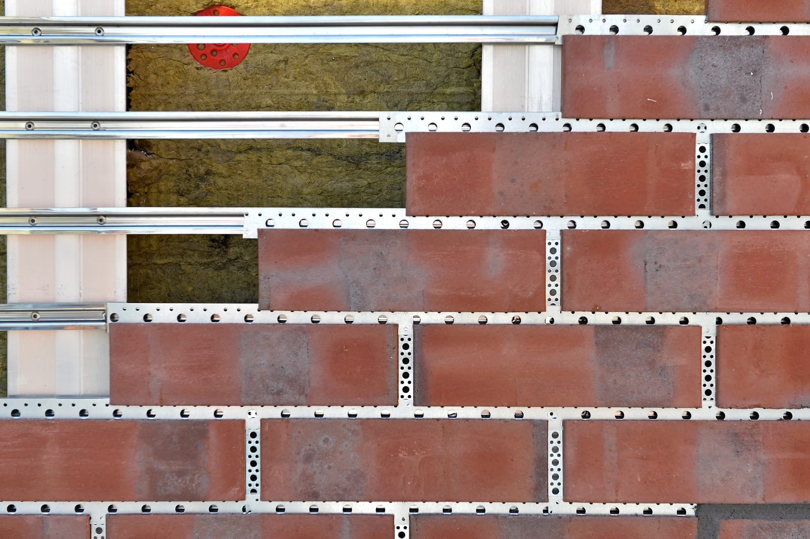 Fachada ventilada en obra: perfiles metálicos horizontales y rieles perforados sobre aislante de lana mineral, con piezas cerámicas rojas en formato de ladrillo colocadas por hiladas