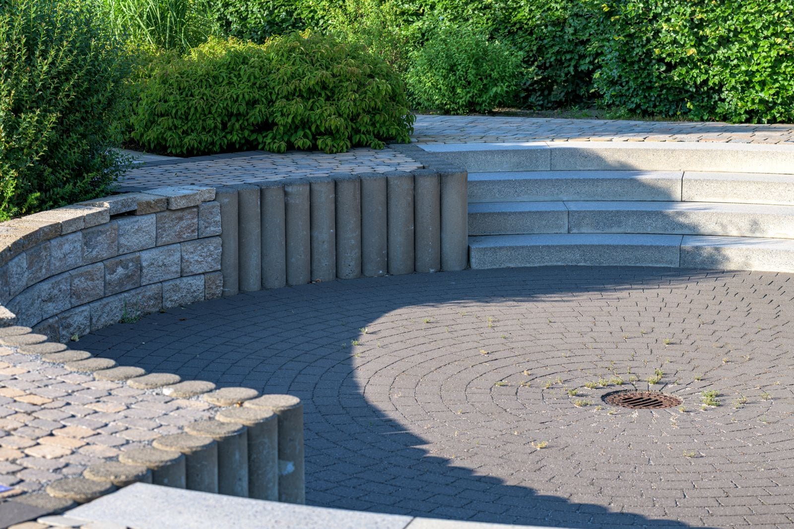 Plaza circular pavimentada con adoquines dispuestos en anillos concéntricos y un sumidero en el centro, rodeada de escalones, un muro curvo de bloques y una hilera de palisadas, con setos y arbustos al fondo