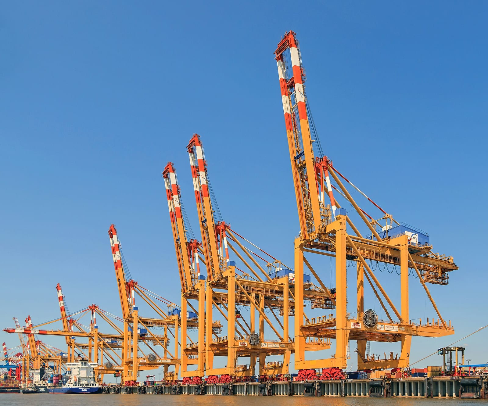 Grúas pórtico amarillas con plumas rojas y blancas alineadas en el muelle de una terminal de contenedores, con un buque atracado a la izquierda y cielo azul despejado