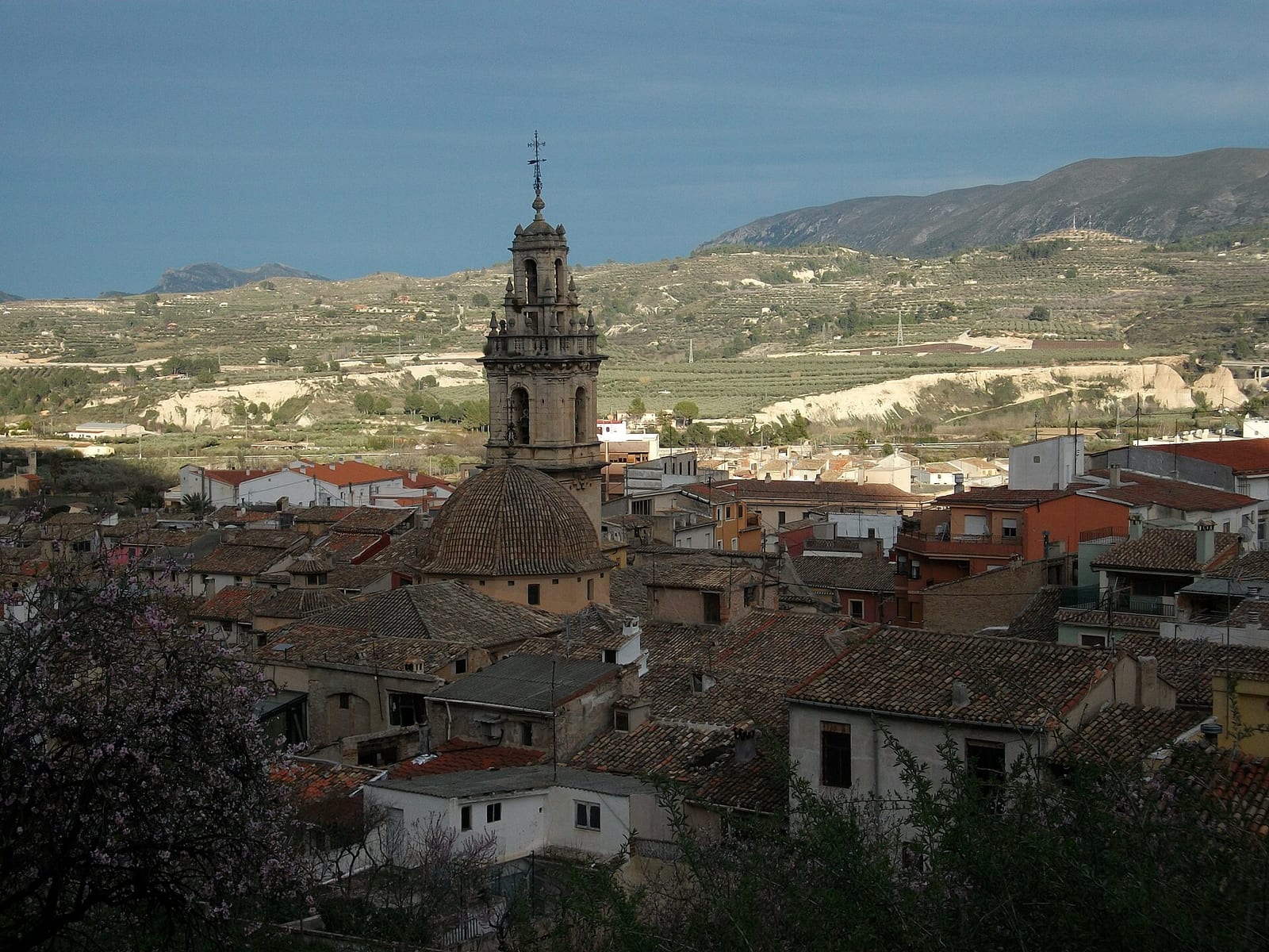 Panorámica de los tejados de teja del casco antiguo de Cocentaina, con la cúpula y la torre campanario de una iglesia en el centro y laderas con bancales y montañas al fondo