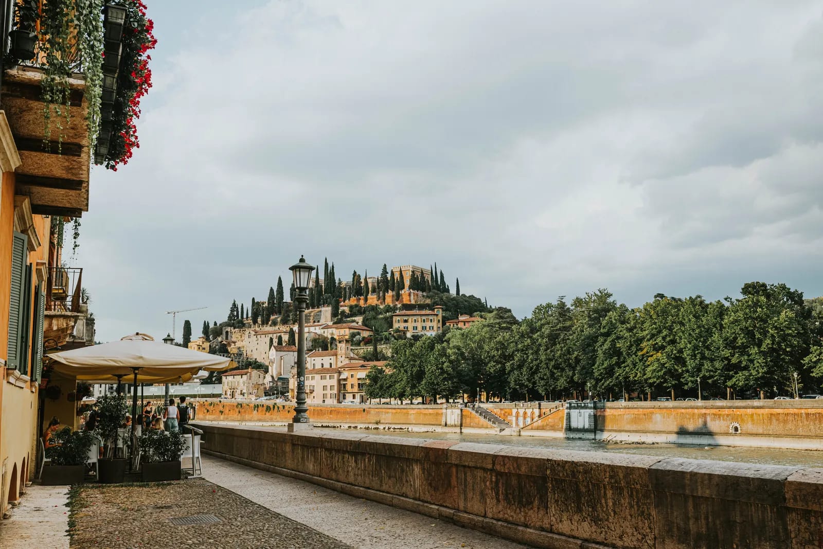 Paseo junto al río Adigio en Verona, con una colina con edificios históricos entre cipreses al fondo, bajo un cielo nublado