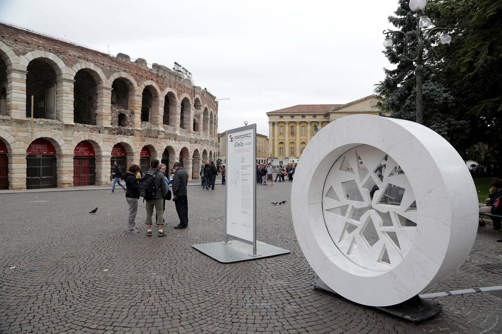Escultura circular de mármol blanco en una plaza junto al anfiteatro romano de Verona, con público observando un panel informativo