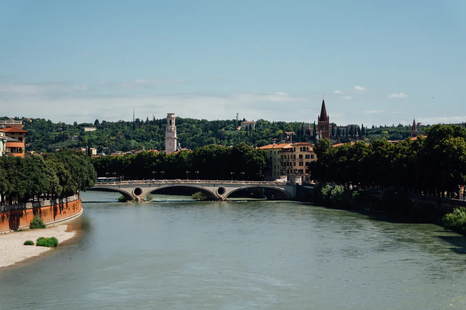 El río Adigio recorre Verona con un puente de piedra y las torres del casco histórico al fondo, bajo un cielo despejado