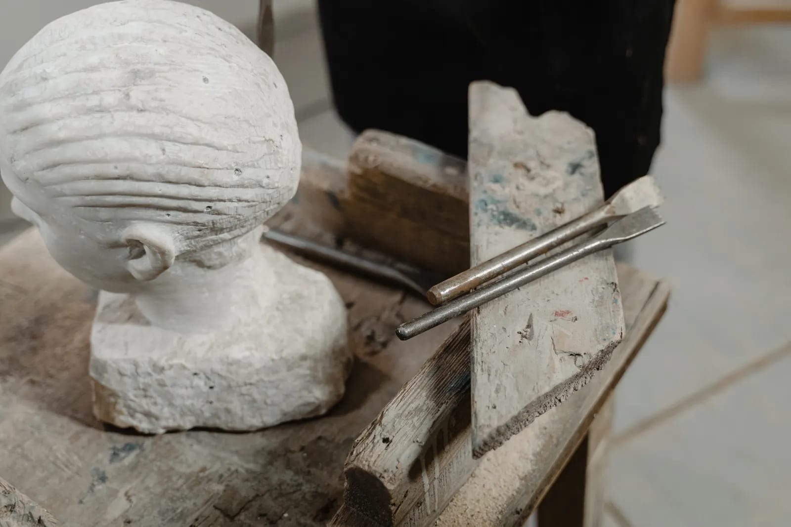 Escultura de una cabeza en piedra clara junto a dos cinceles de talla sobre un banco de madera en un taller