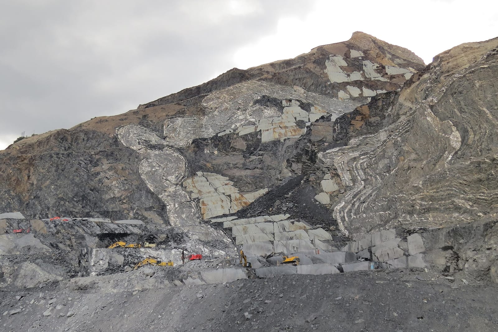 Frente de una cantera de pizarra en ladera de montaña, con capas de roca gris oscuro plegada y excavadoras trabajando al pie del talud