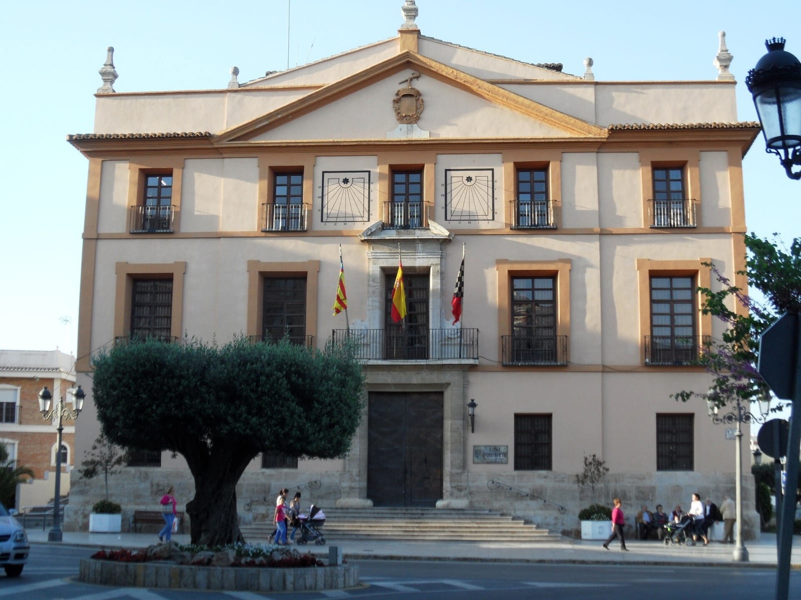 Fachada de un edificio consistorial de estilo clásico con balcones, escudo en el frontón y peatones cruzando la plaza de acceso