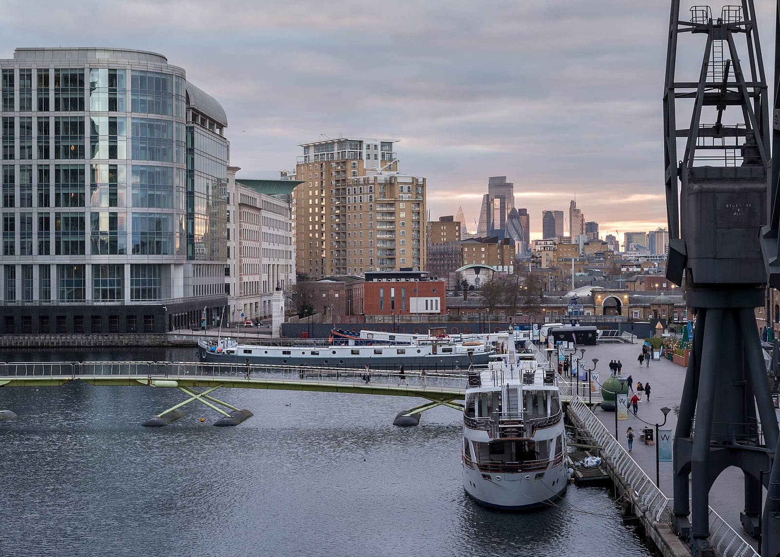 Dársena de Canary Wharf con barcos atracados, una pasarela sobre el agua y una grúa portuaria en primer plano; al fondo, los rascacielos de la City de Londres al atardecer