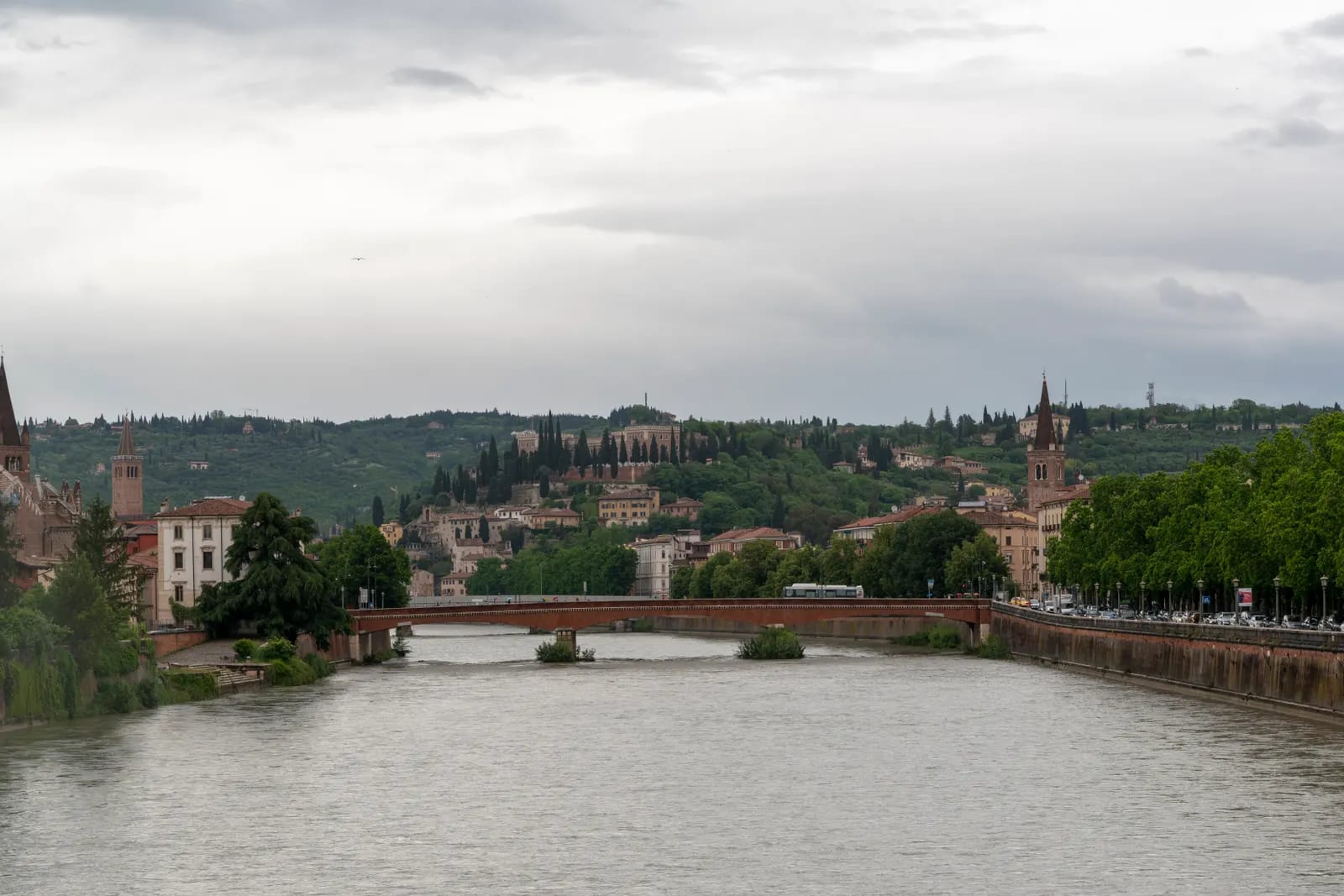Vista del río Adige a su paso por Verona, con un puente de ladrillo, campanarios y una colina de edificios históricos y cipreses bajo un cielo nublado