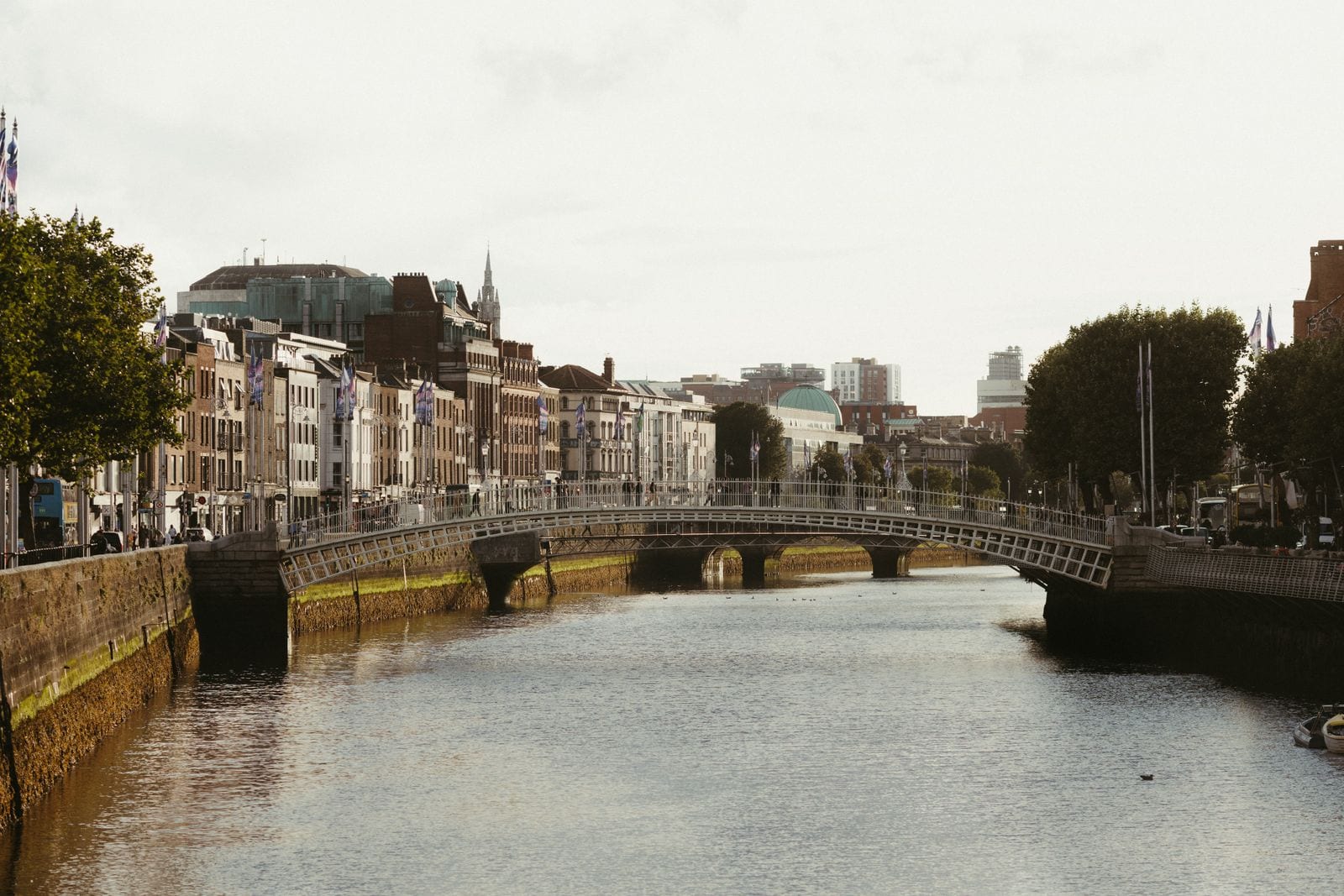 Puente peatonal blanco de hierro sobre el río Liffey, con edificios de ladrillo y piedra en el muelle y cielo nublado, en Dublín