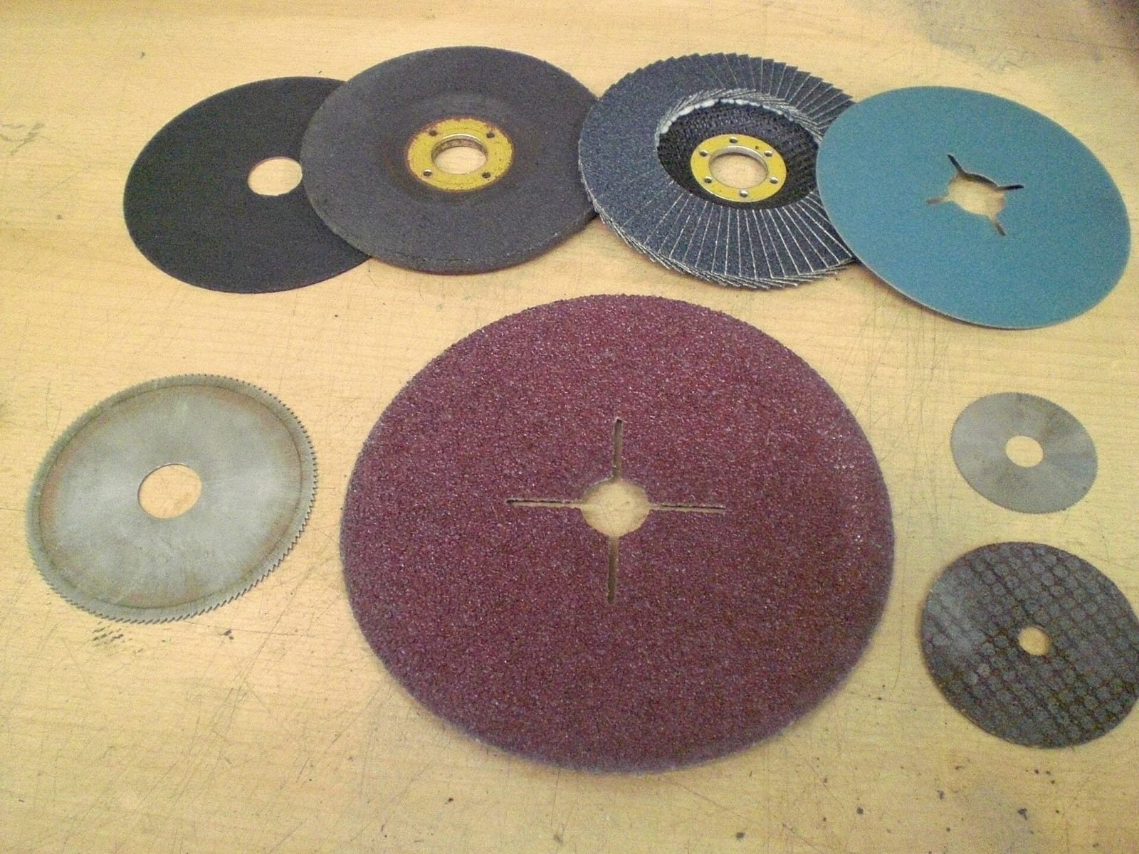 Surtido de discos para amoladora sobre una mesa de madera: un disco de láminas gris azulado, discos de fibra abrasiva granate y turquesa, discos de desbaste y de corte y dos discos de sierra metálicos
