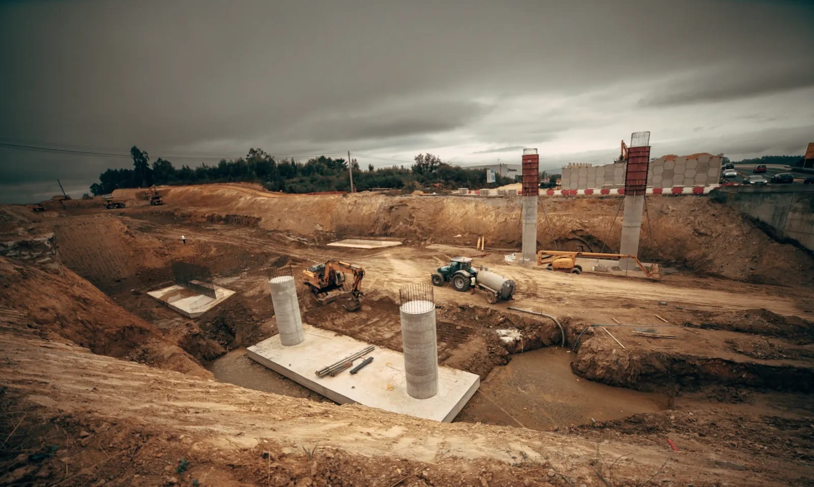 Obra de excavación con maquinaria, pilares de hormigón y cimentación sobre terreno arcilloso bajo un cielo nublado
