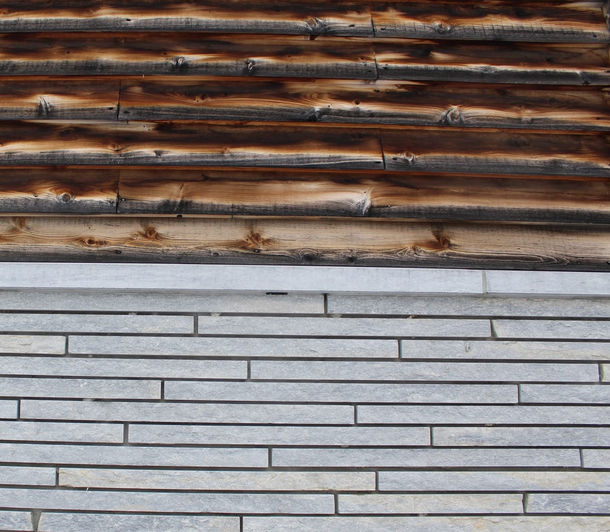 Fachada de un edificio con franjas de madera quemada en la parte superior y revestimiento de listones de caliza gris en la inferior
