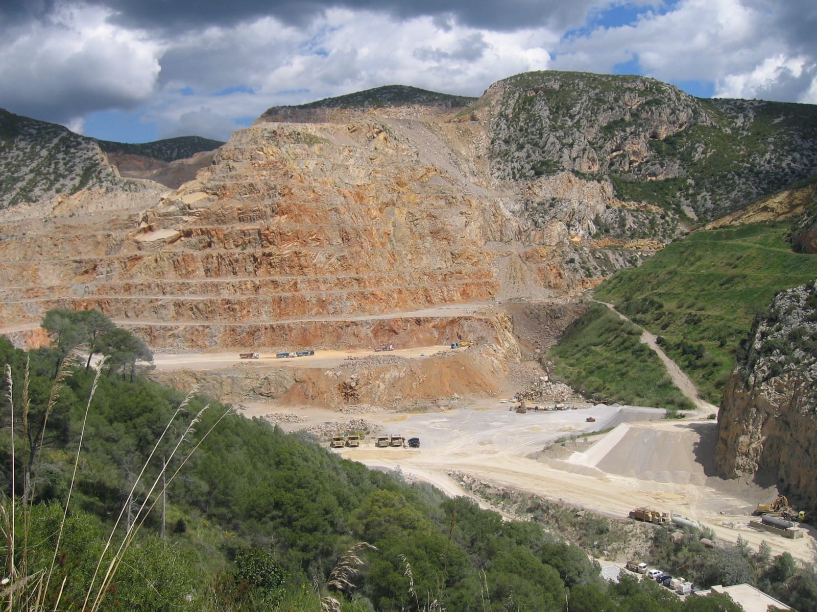 Gran cantera de caliza a cielo abierto en un macizo montañoso, con bancos escalonados de roca ocre y maquinaria en la explanada inferior