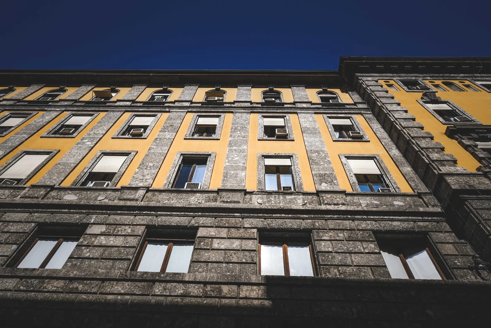 Fachada de un edificio histórico vista desde abajo, con sillares de piedra gris en la base y los recercados, y paños amarillos, bajo un cielo azul intenso