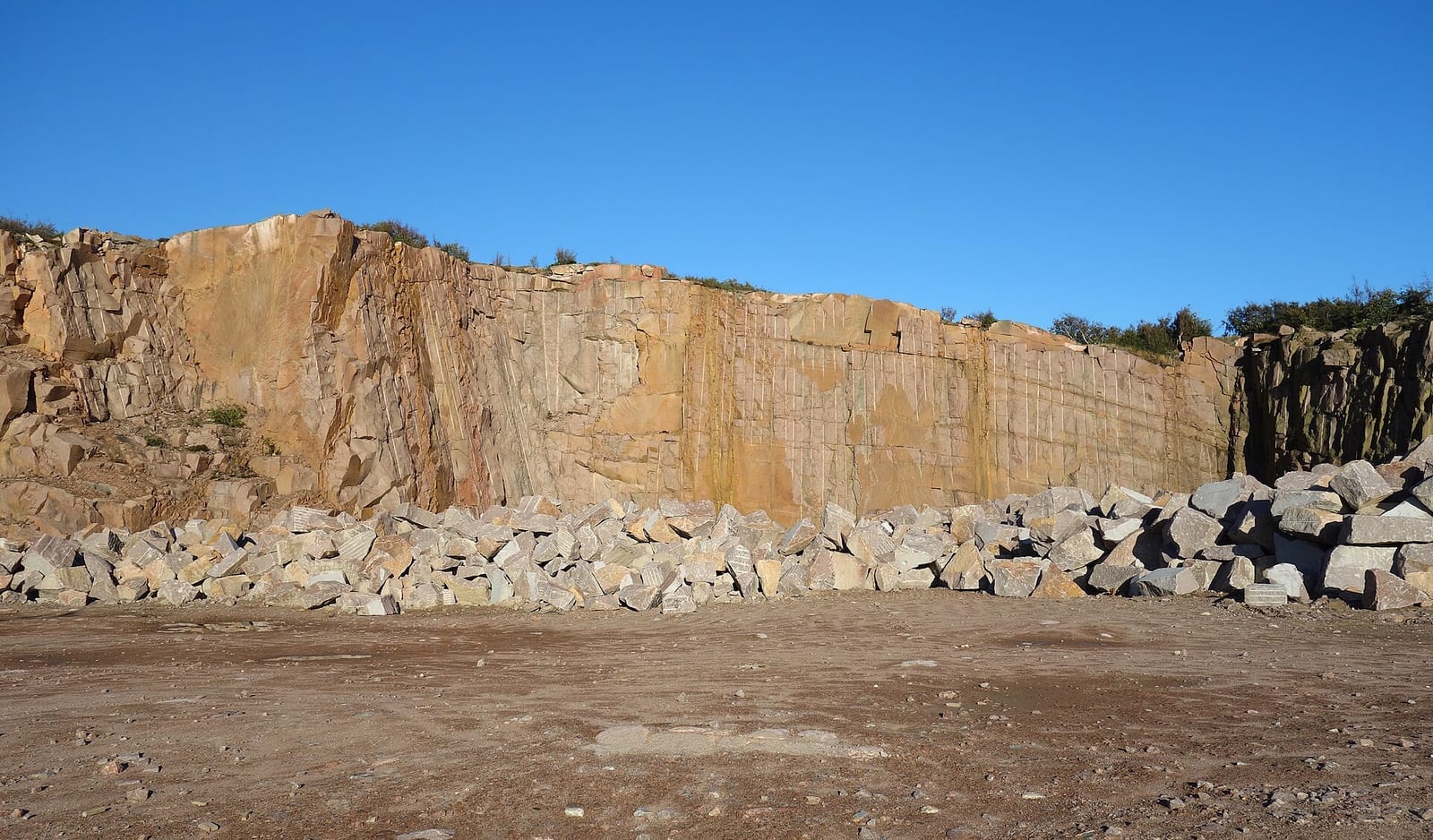 Frente de cantera de granito de tonos anaranjados, con marcas verticales de perforación y bloques de piedra amontonados al pie, bajo un cielo azul despejado