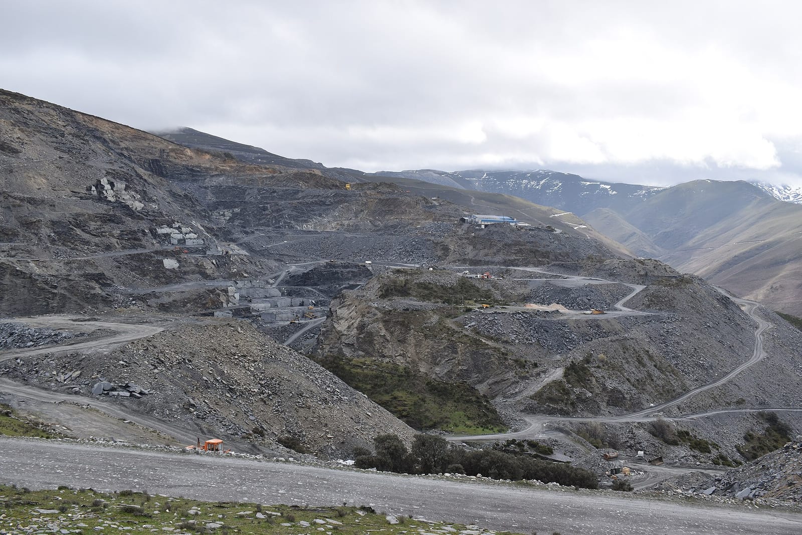 Explotación minera a cielo abierto en una ladera de montaña, con pistas, escombreras de roca oscura y cumbres con nieve al fondo