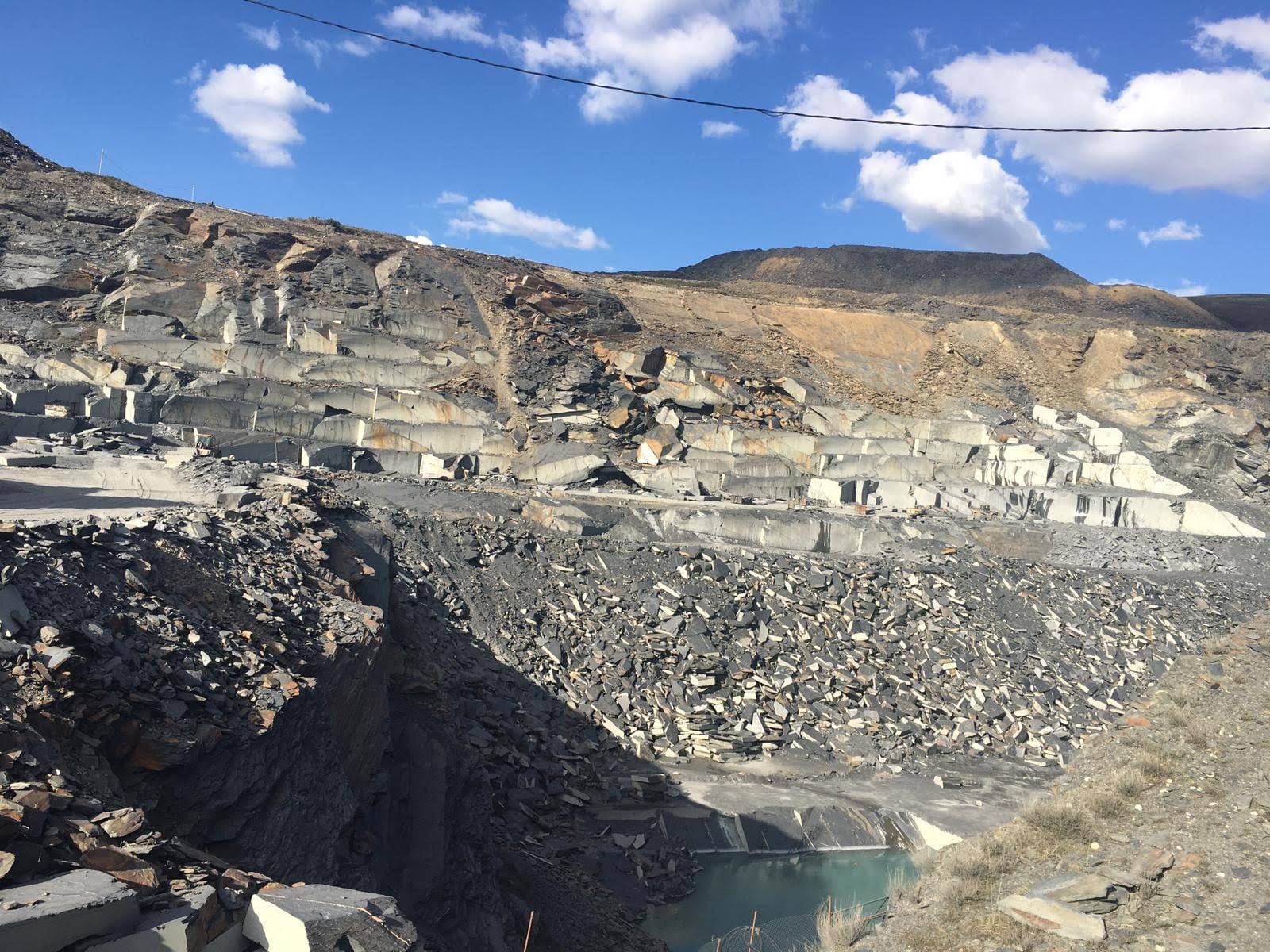Cantera de pizarra a cielo abierto con bancos de roca gris, escombrera de lajas en primer plano y una balsa de agua turquesa