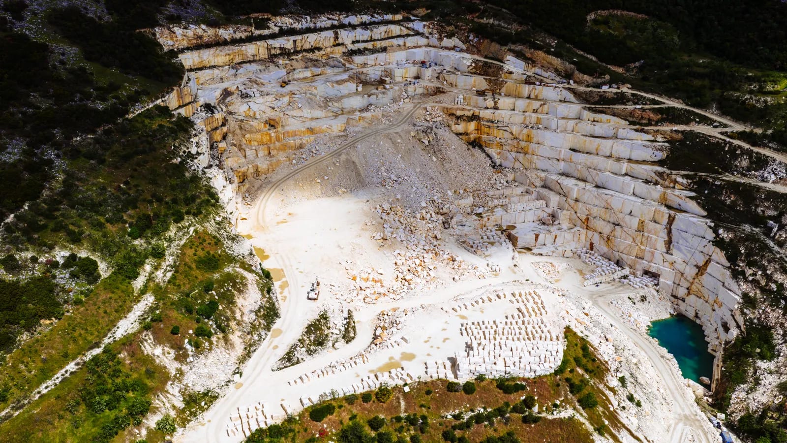 Vista aérea de una cantera de mármol a cielo abierto con bancales escalonados, bloques cortados apilados y una balsa de agua turquesa al fondo