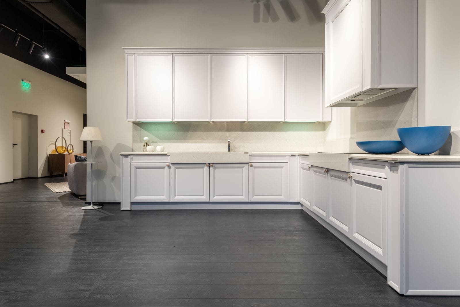 Cocina en L de armarios blancos con encimera y frontal de tono claro y dos cuencos azules, montada en una sala de exposición amplia con suelo oscuro y una lámpara de pie al fondo