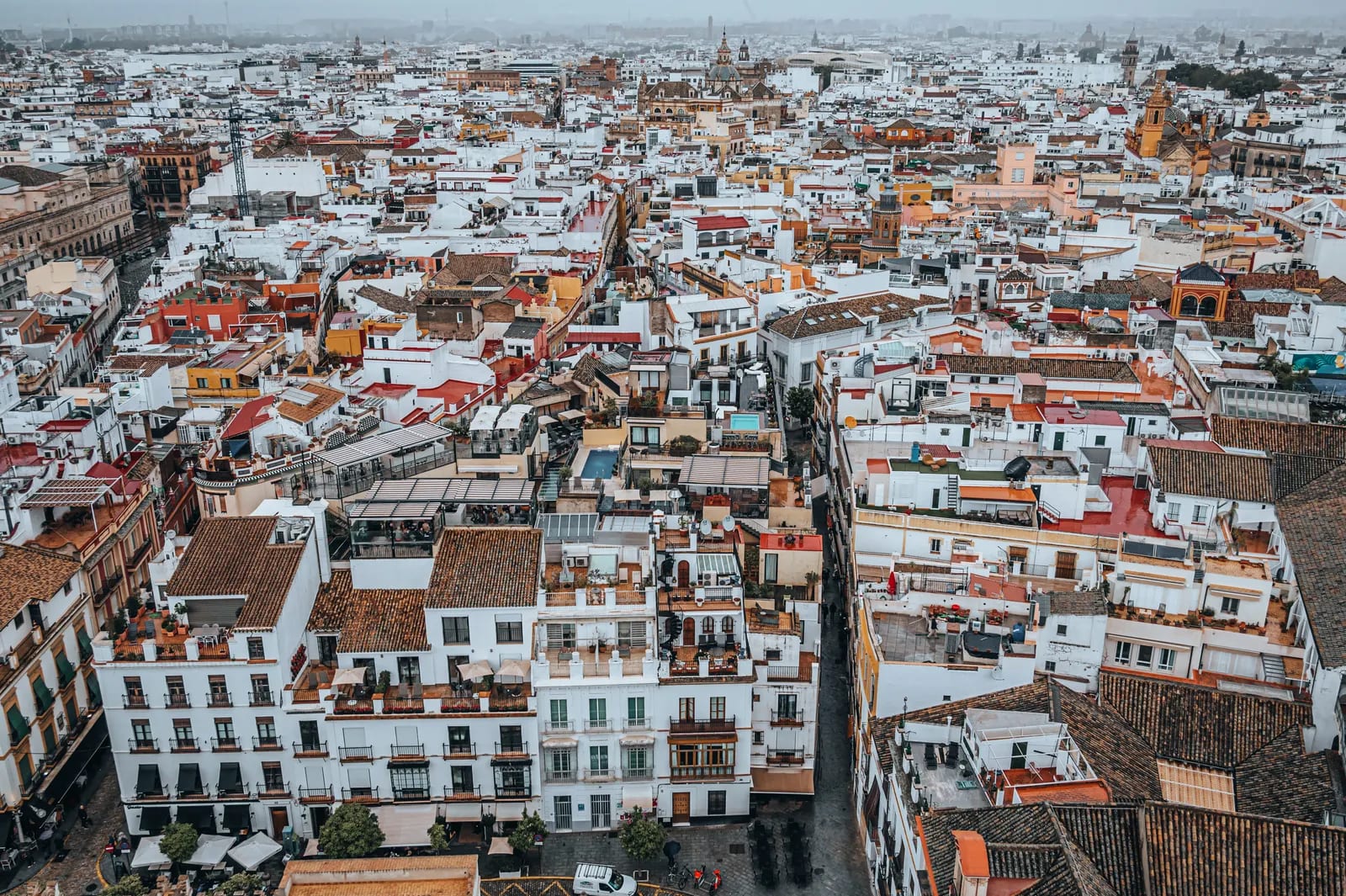 Vista aérea de los tejados y las calles de Sevilla, con edificios blancos y ocres y torres al fondo, bajo un cielo nublado