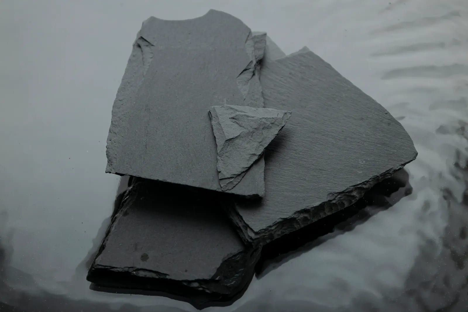 Varias lajas de pizarra gris oscuro de bordes irregulares apiladas, con un fragmento triangular encima, sobre una superficie oscura y brillante