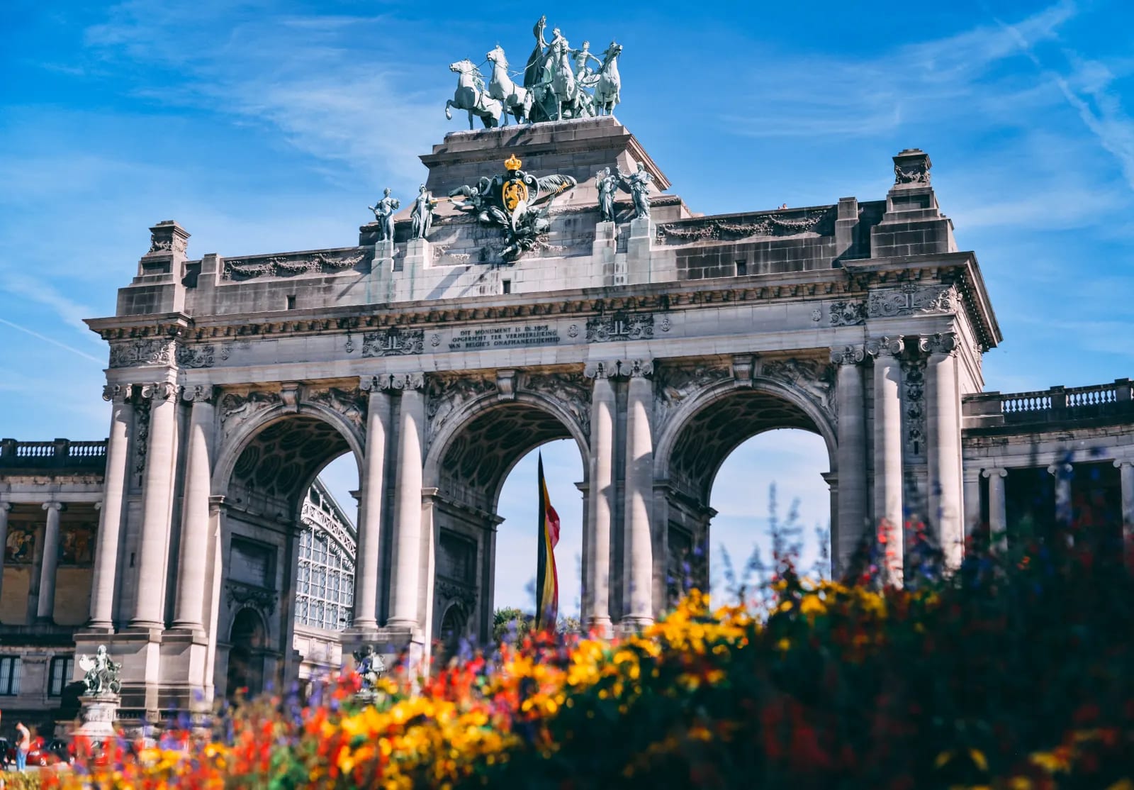 Arco monumental de piedra con esculturas y cuadriga en lo alto, bandera belga entre los arcos y flores de colores en primer plano
