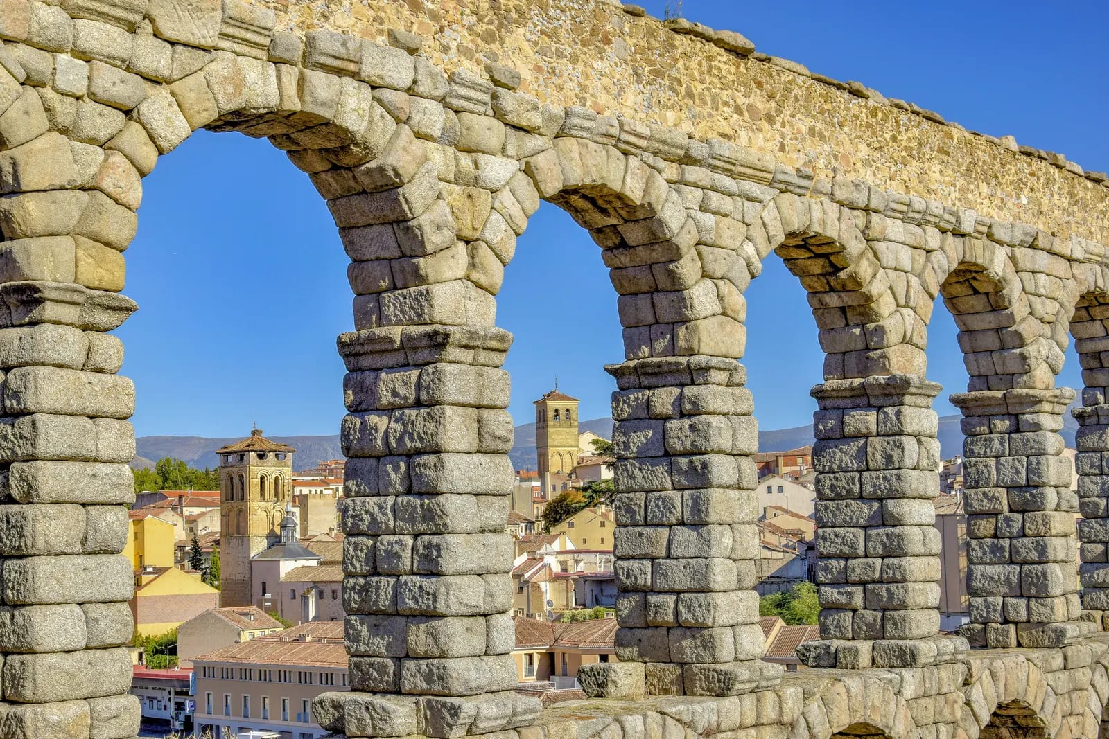 Arcos de sillería de granito del acueducto romano con tejados y torres de Segovia al fondo bajo un cielo azul despejado