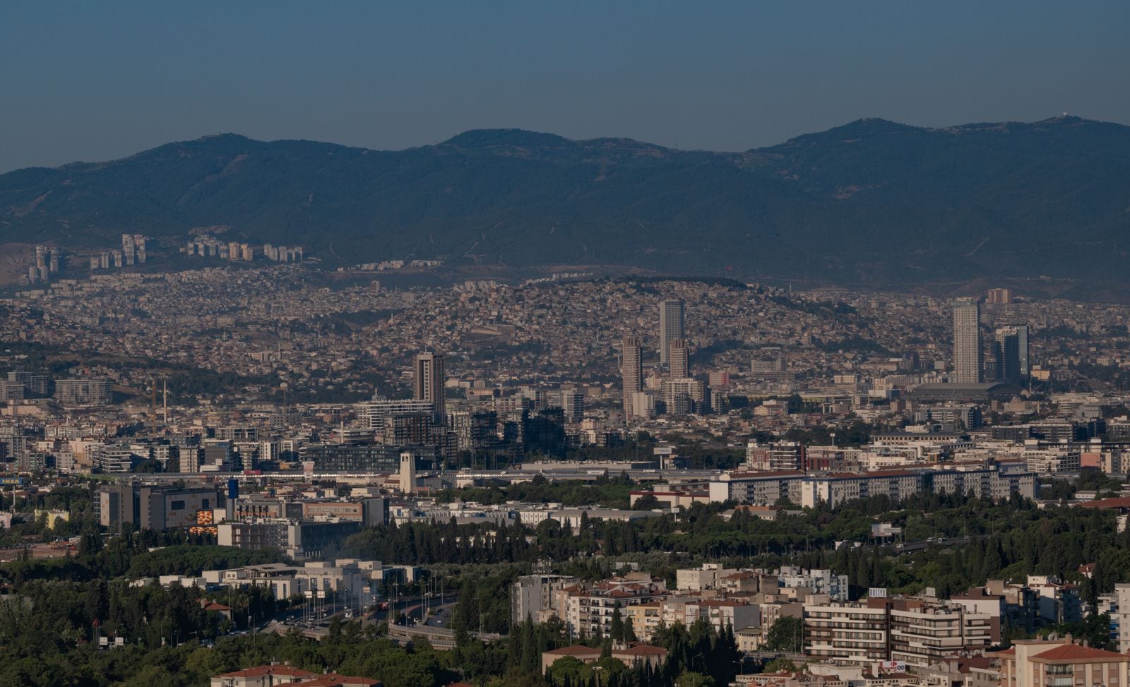 Vista elevada de la ciudad de Izmir con torres y bloques de viviendas que suben por las laderas, montañas al fondo y cielo azul con algo de bruma