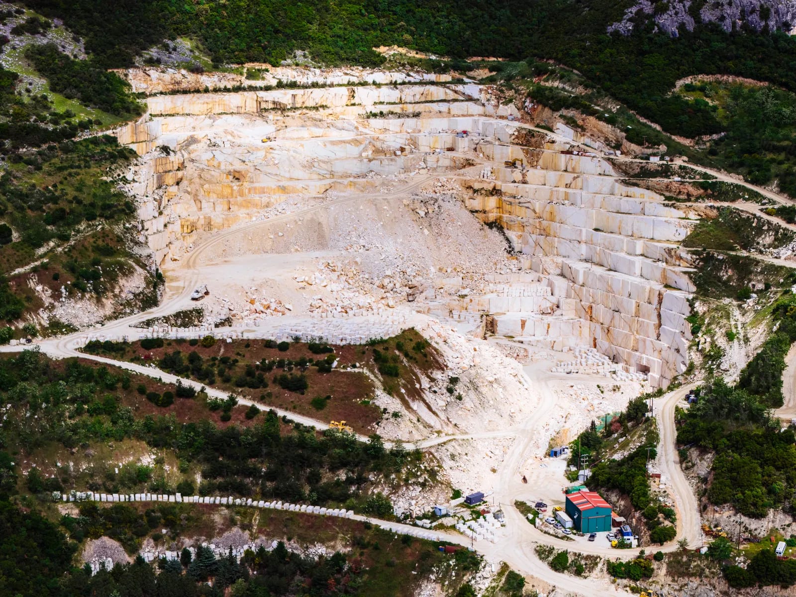 Vista aérea de una cantera de mármol en bancos escalonados rodeada de bosque, con pistas de tierra y una nave verde
