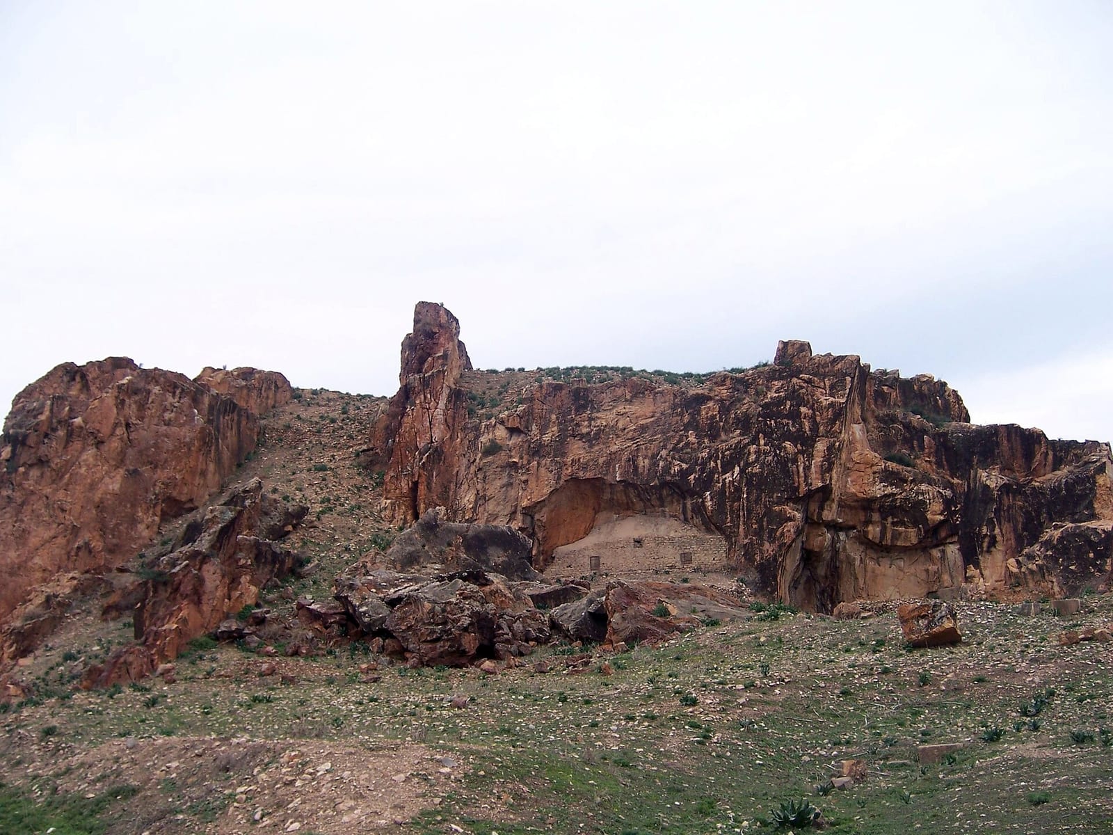 Frente rocoso rojizo de una antigua cantera romana de mármol sobre una ladera de hierba y piedras bajo un cielo nublado