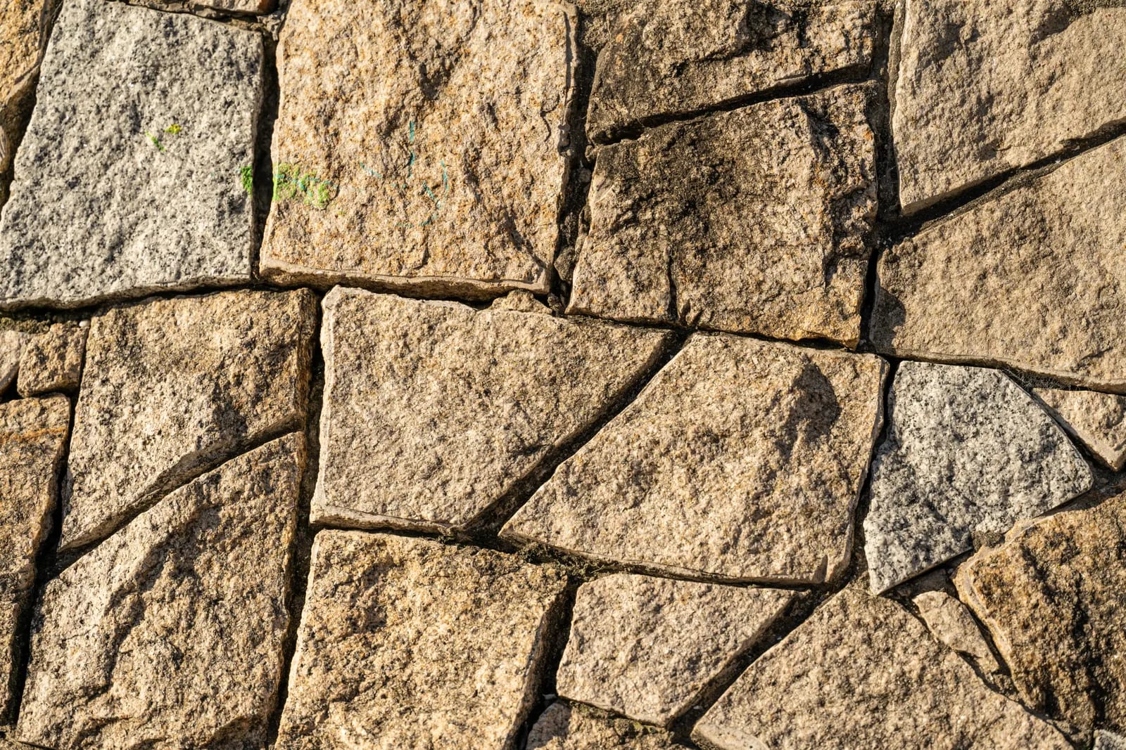 Piezas irregulares de piedra granítica de tonos grises y dorados, con grano punteado y juntas oscuras, bajo luz cálida