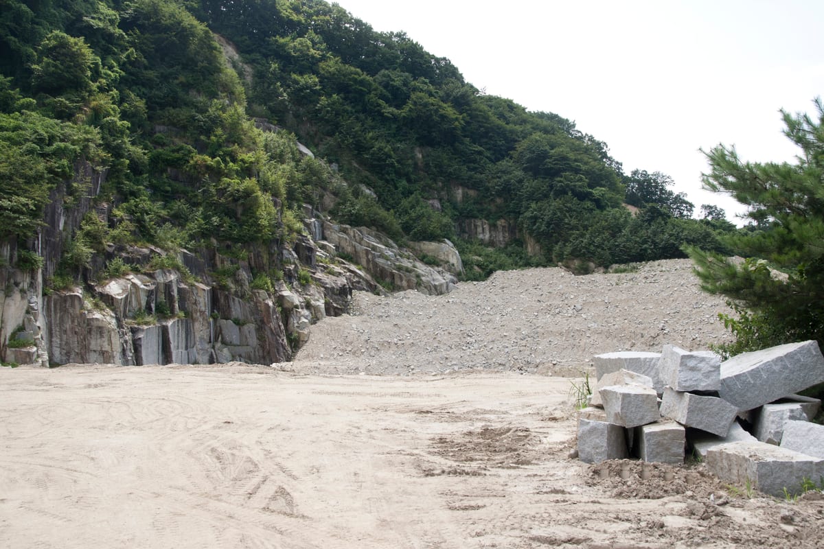 Frente de cantera de granito en una ladera boscosa con bloques escuadrados apilados junto a una explanada de arena