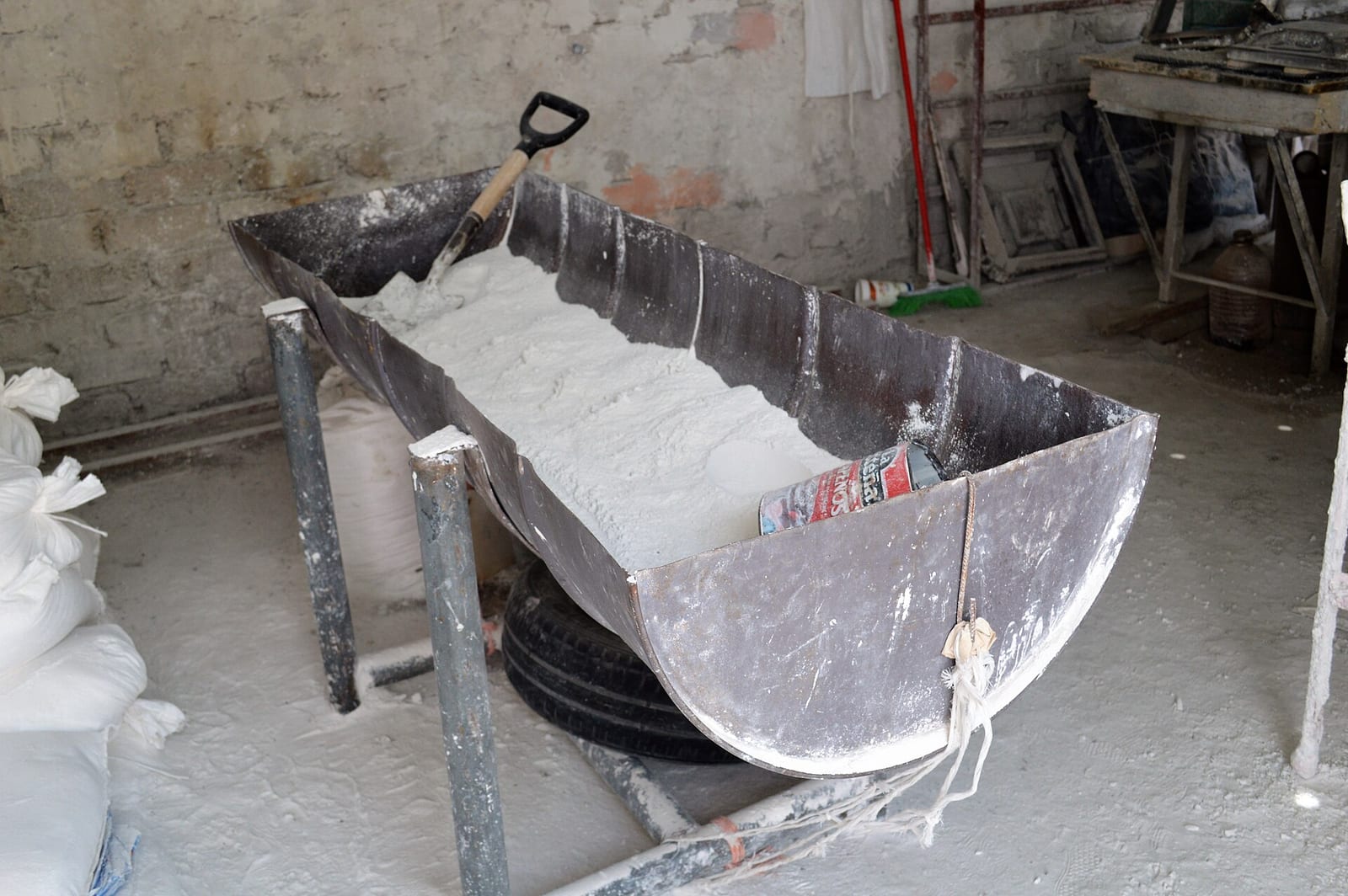 Artesa metálica llena de polvo blanco de mármol con una pala dentro, en un taller con suelo y sacos cubiertos de polvo