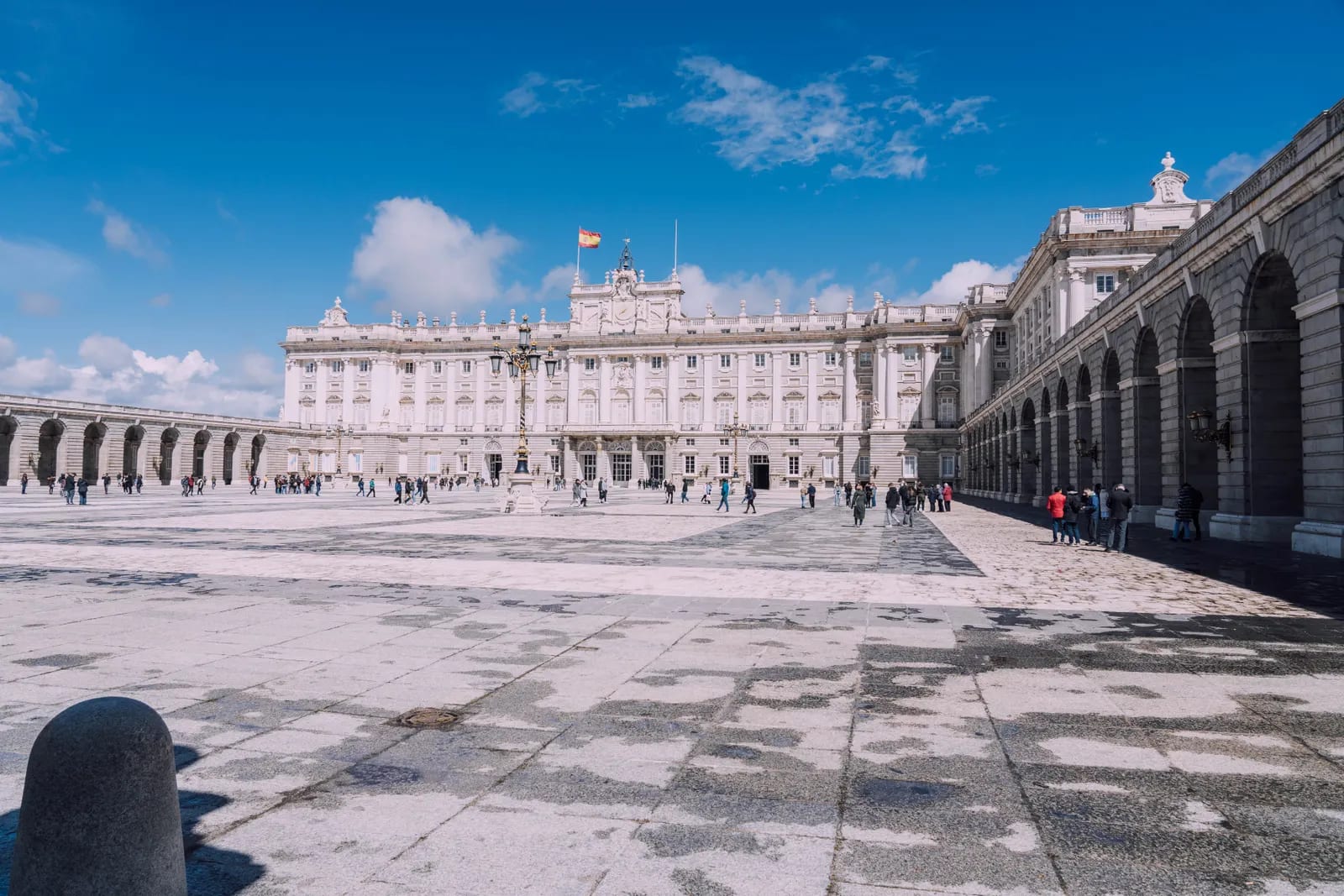 Fachada de piedra clara del Palacio Real de Madrid y su plaza empedrada, bajo un cielo azul con nubes