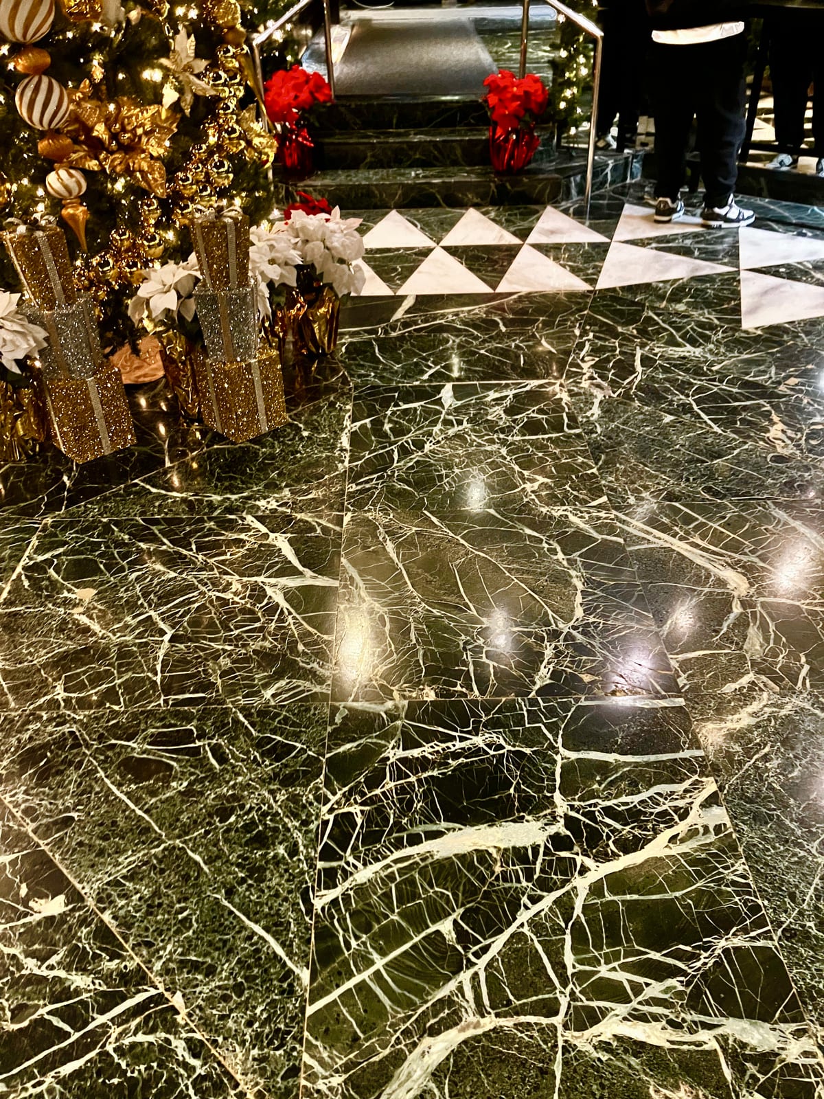Suelo pulido de mármol verde oscuro con vetas blancas en un vestíbulo de hotel, con un árbol de Navidad a un lado y una franja de triángulos claros ante unos escalones al fondo