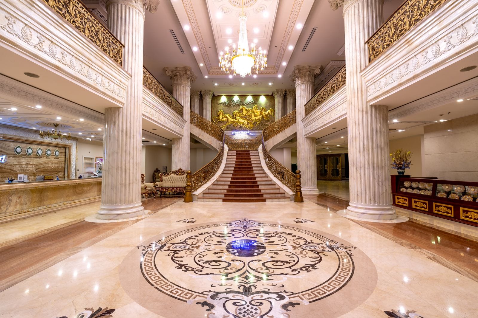 Vestíbulo de hotel con doble escalera, columnas acanaladas de piedra clara y suelo pulido con un gran medallón de mármoles de varios colores