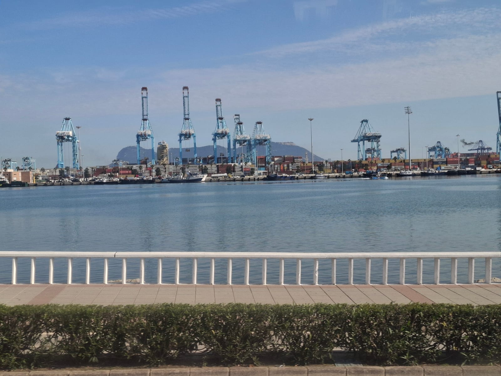Grúas pórtico azules de la terminal de contenedores del puerto de Algeciras, al otro lado de la bahía, con el Peñón de Gibraltar al fondo y un paseo marítimo en primer plano