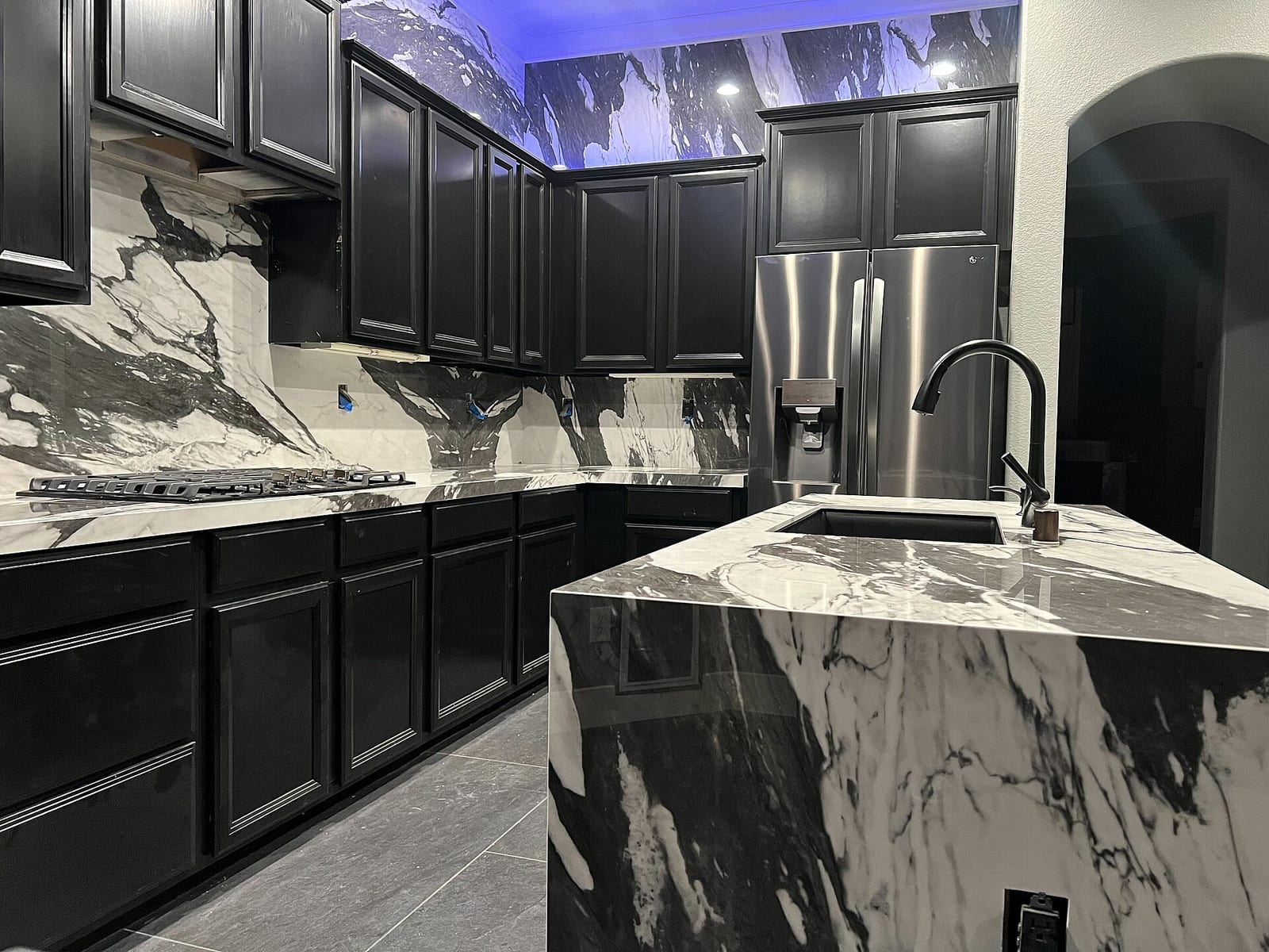 Cocina con muebles negros, encimera y frontal blancos con vetas negras y grises, e isla con canto en cascada del mismo material y fregadero bajo encimera