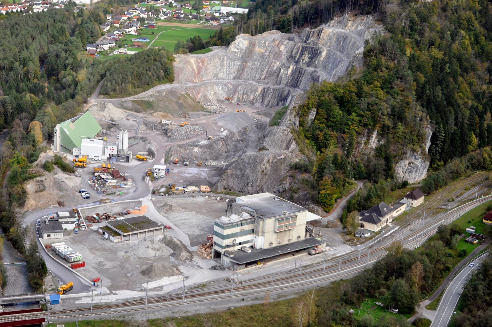 Vista aérea de una cantera escalonada en una ladera boscosa junto a una planta de cemento con silos, camiones hormigonera y una vía de tren