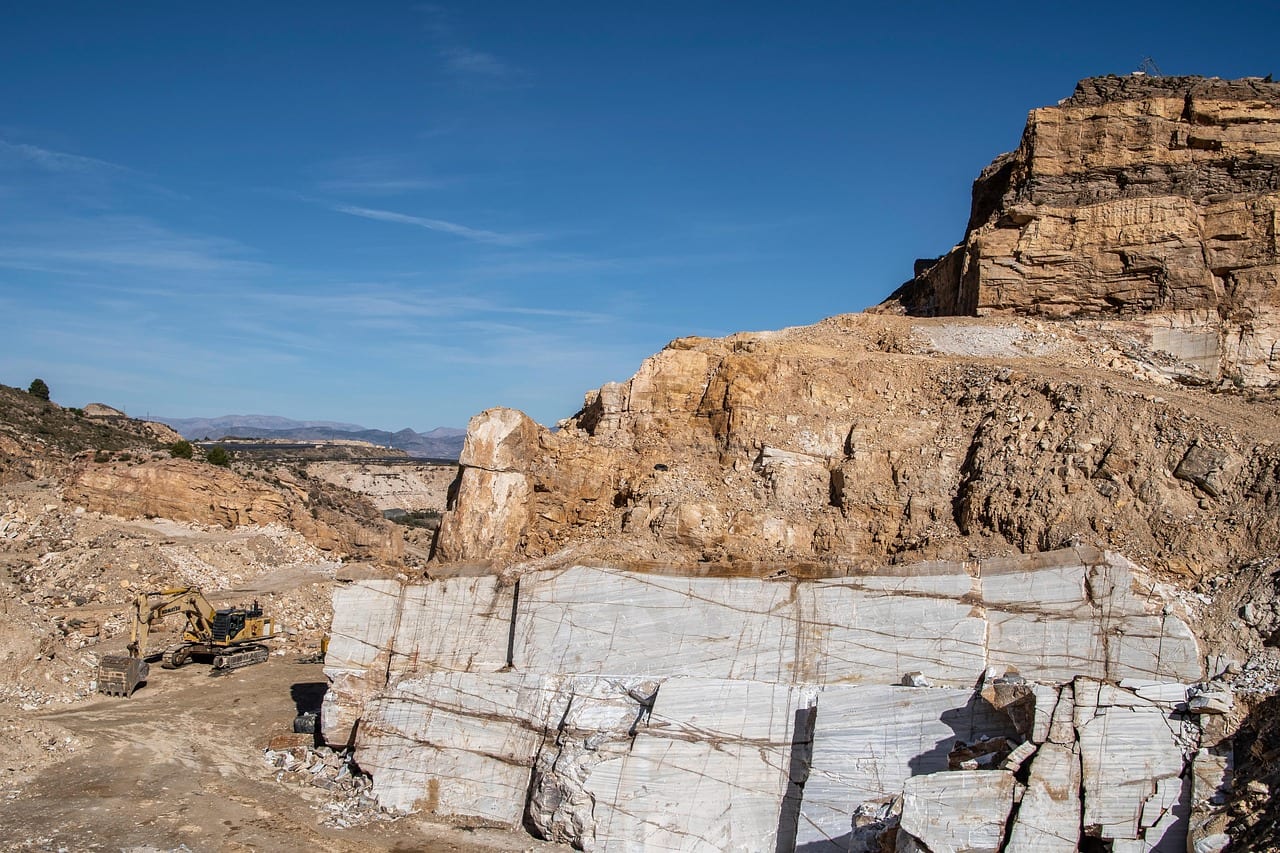 Frente de cantera de mármol blanco con grandes bloques fracturados en primer plano, una excavadora al fondo y cielo azul despejado