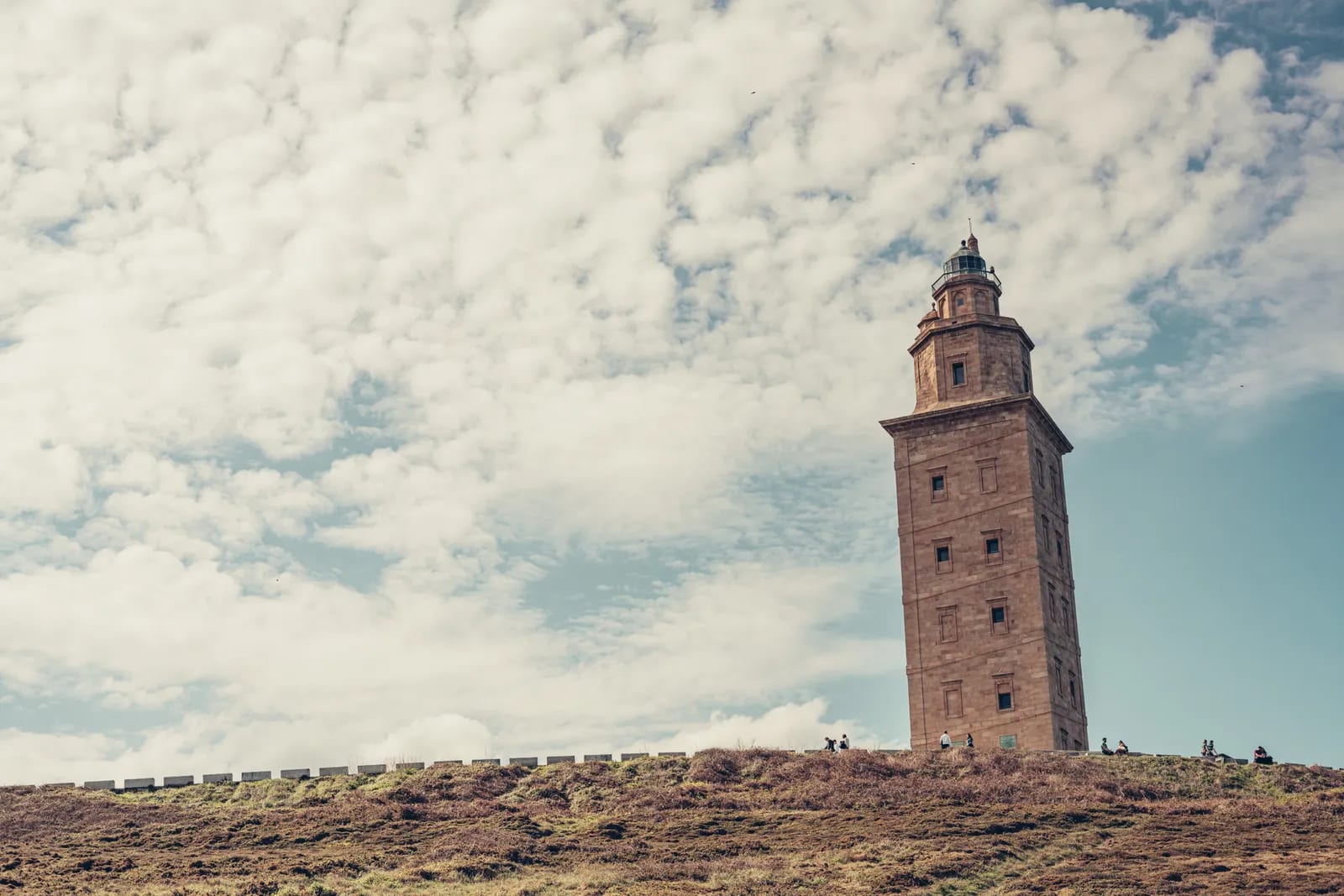 Torre de Hércules vista desde abajo sobre una loma de brezo, con algunos visitantes pequeños al pie y un cielo cubierto de nubes blancas con claros azules