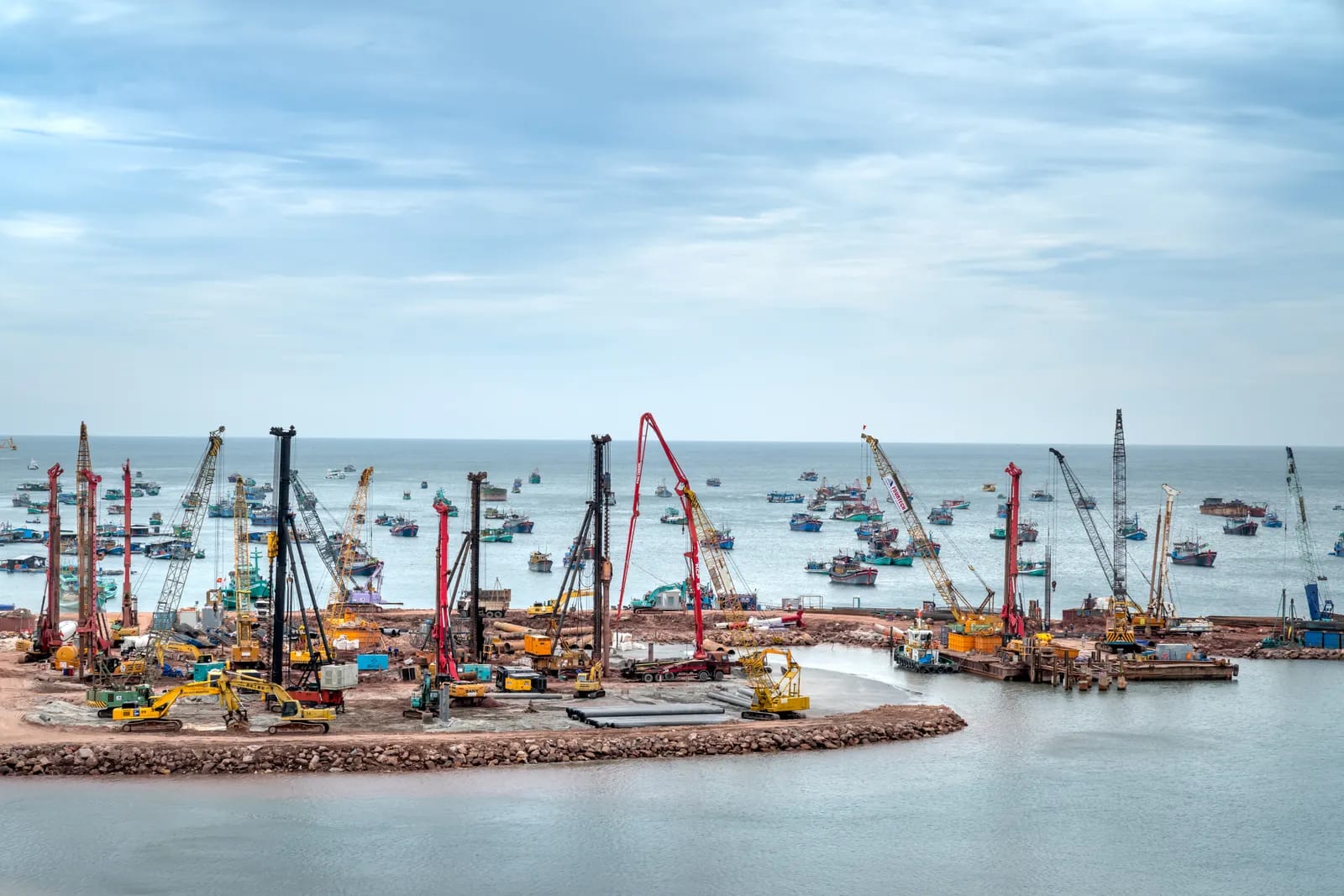 Obra costera con grúas, pilotadoras y excavadoras amarillas sobre una explanada junto al mar, bajo un cielo nublado