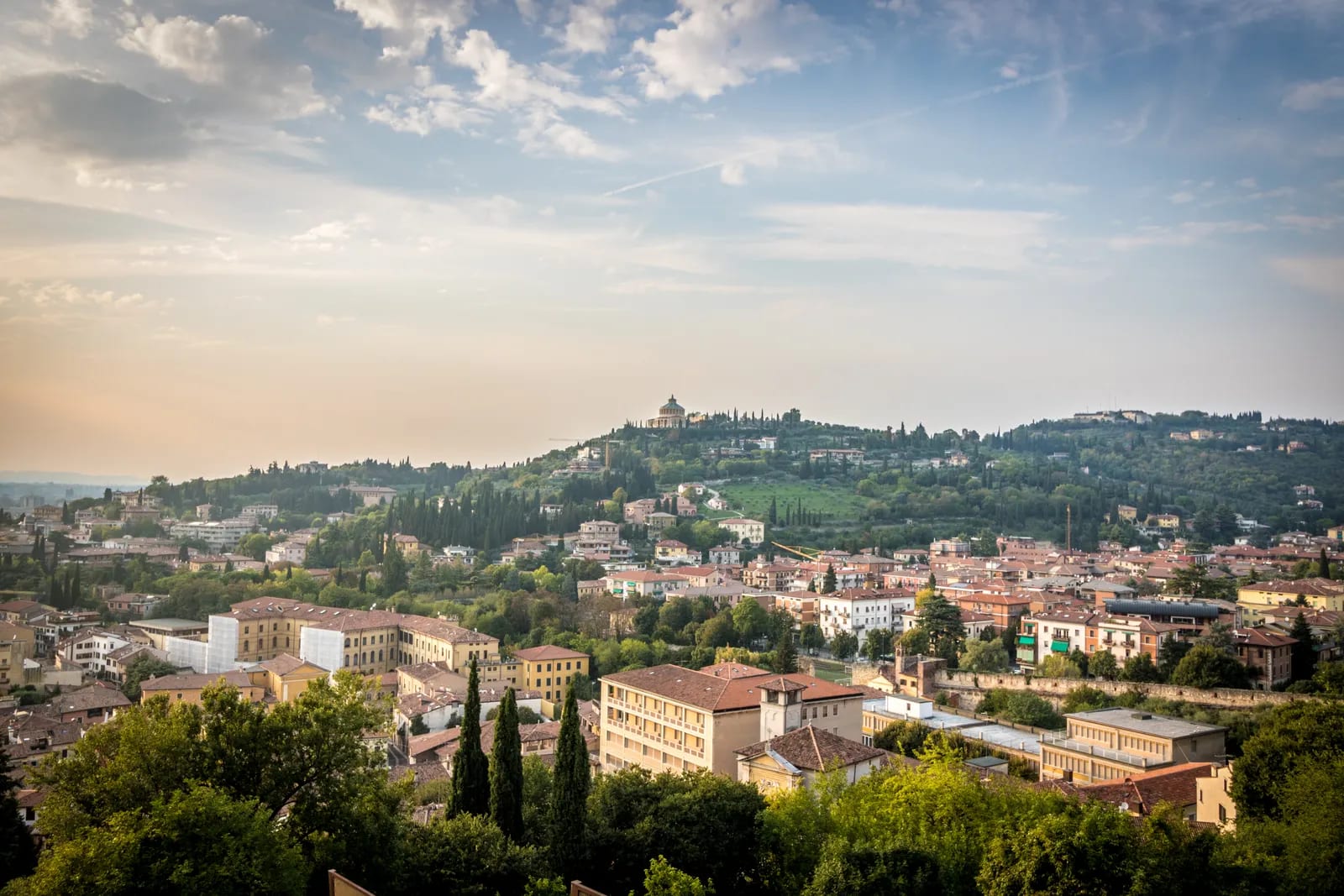 Vista elevada de Verona con tejados de teja, cipreses y una colina arbolada coronada por un santuario bajo un cielo con nubes
